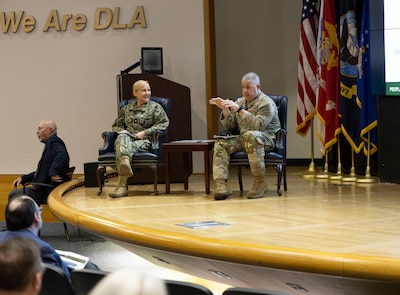 A man and a woman are seated on stage in an auditorium. We Are DLA is emblazoned on the wall behind them and their are military flags adjacent to them. She is a blond and is wearing a Navy utility uniform and he is bald and is wearing an Air Force OCP uniform.