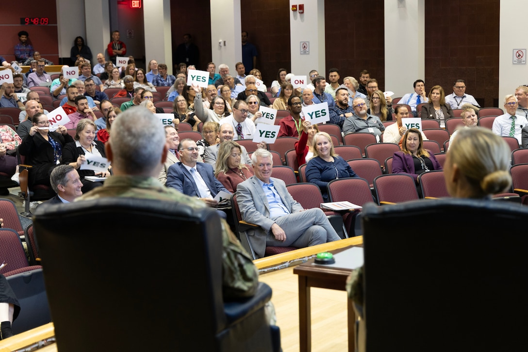 Two people sit on stage. Both are in military uniforms. One is a man and the other a blond-haired woman. They have buzzers before them.