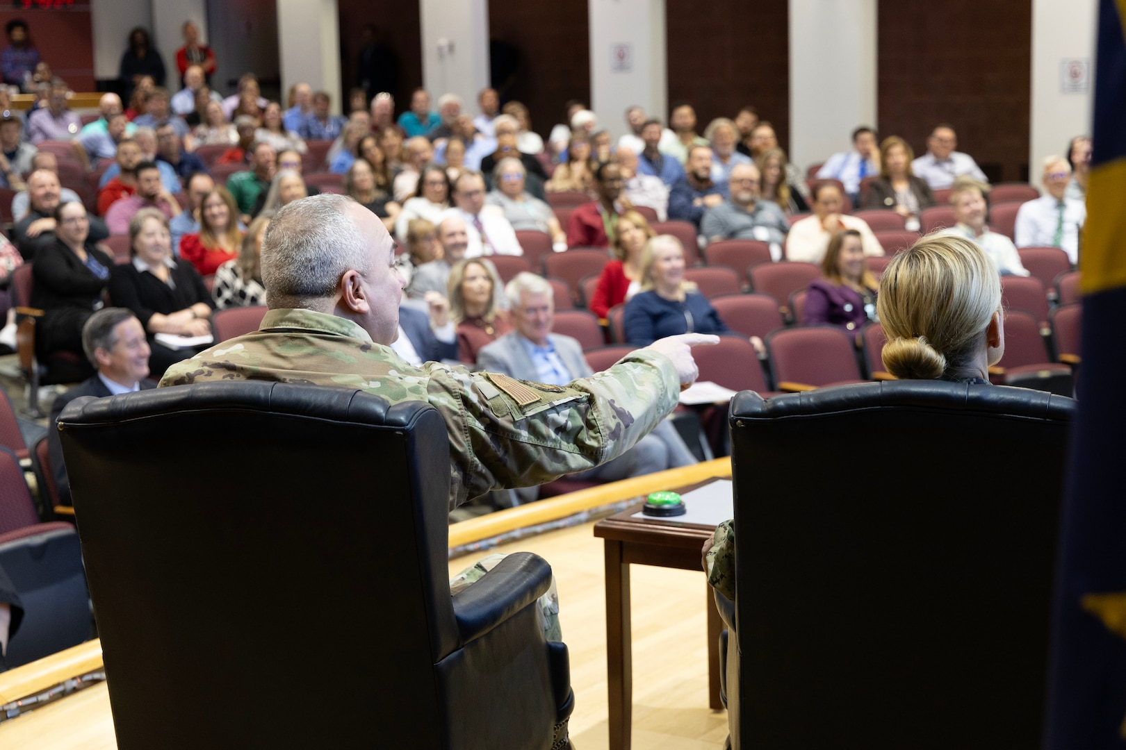 Two people sit on stage. Both are in military uniforms. One is a man and the other a blond-haired woman. They have buzzers before them.