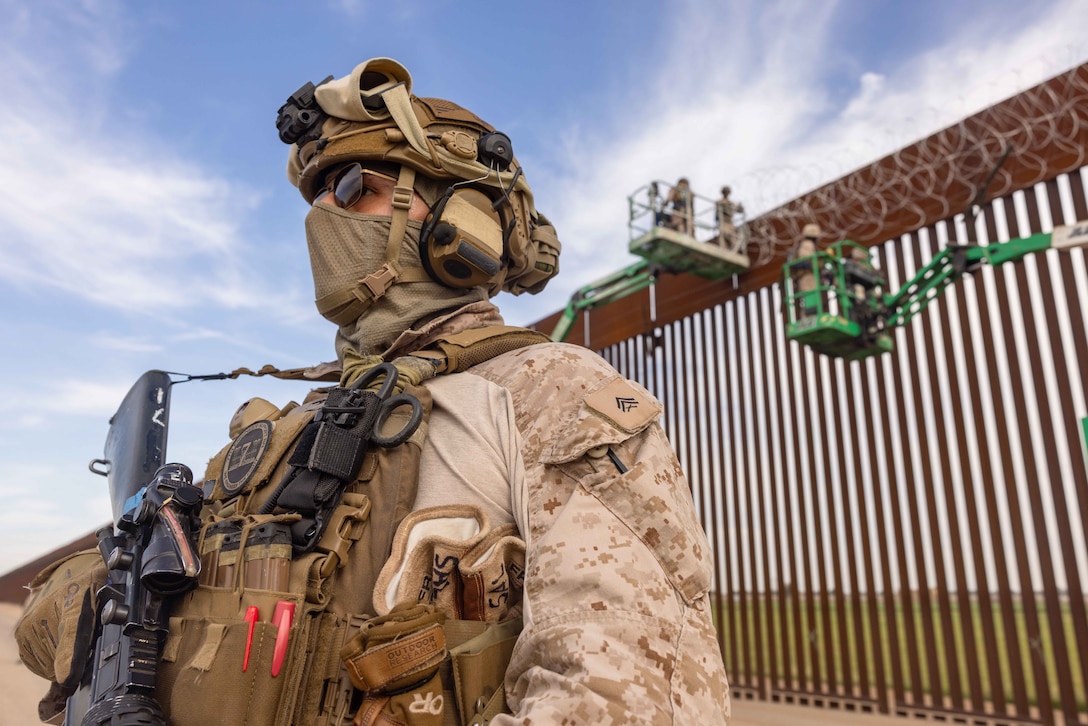A Marine wearing a helmet and a face mask stands watch while Marines on lifts add wire to a large metal fence in a desert-like area.