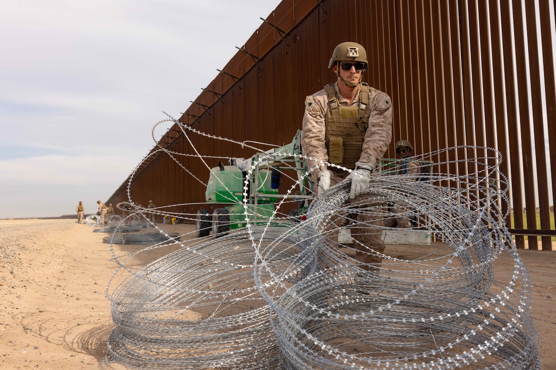 A Marine drags concertina wire near a large metal fence in a desert-like area.