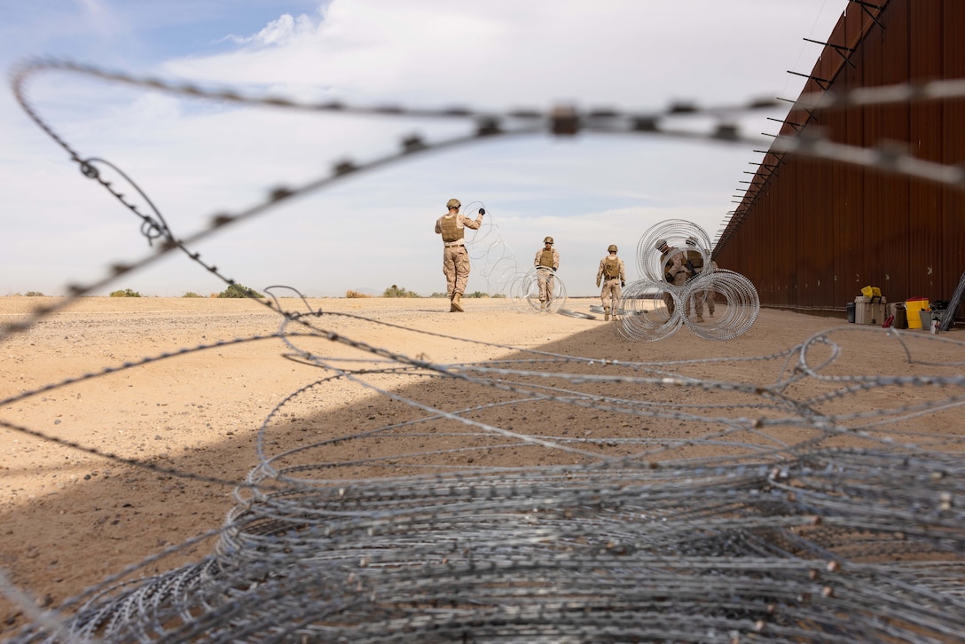 Concertina wire sits in the foreground as Marines work on wire in the distance next to a tall metal fence in a desert-like area.