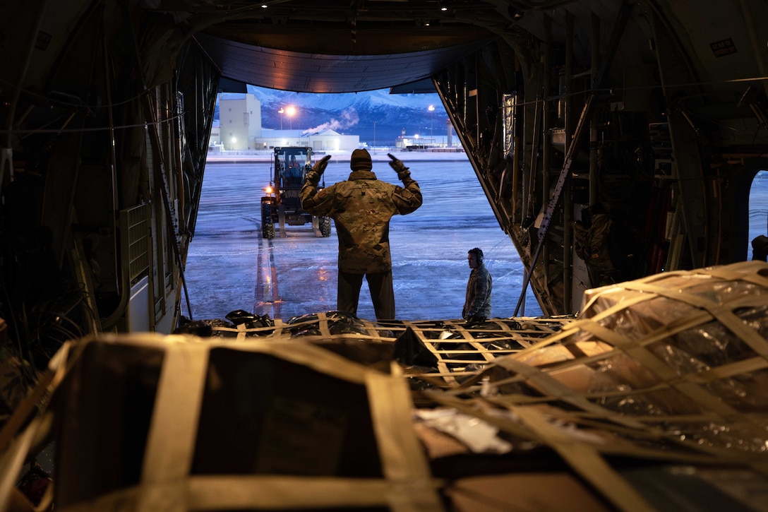 A guardsman uses both hands to direct an oncoming tractor toward the open cargo bay of an aircraft as another guardsman stands outside in icy, dusk-like conditions.