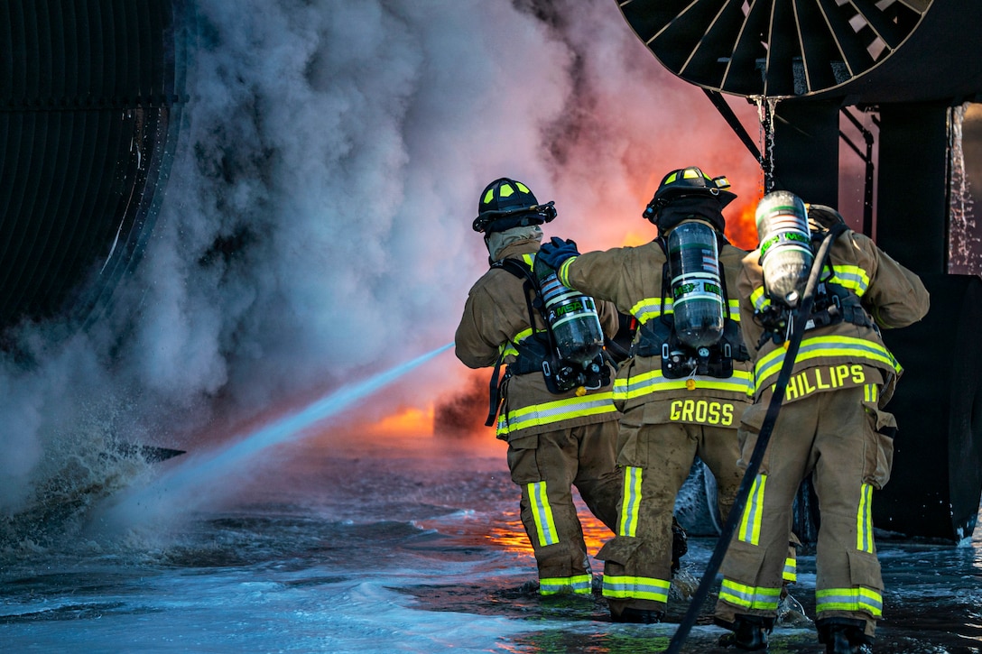Three firefighters wearing helmets and oxygen tanks spray water from a hose toward flames near large equipment outdoors.