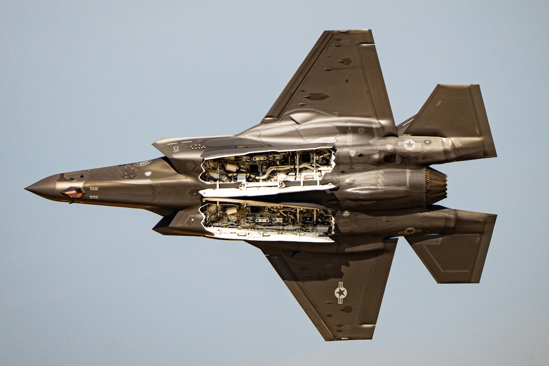 The underside of a fighter jet is seen as it flies in a cloudless sky during the day.