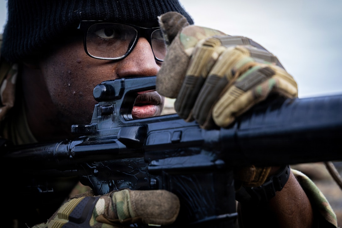 An Army ROTC cadet wearing a black, knit cap and camouflage gloves stares ahead while holding a rifle outdoors during daytime.