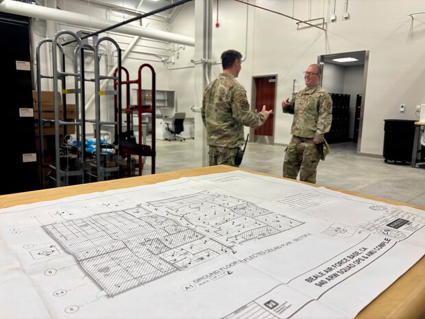 Two airmen talk inside a hangar as blueprints of the new Squadron Operations and Aircraft Maintenance Unit facility sit nearby at Beale Air Force Base, California on Nov. 14, 2025.