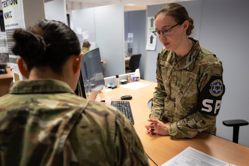 An airman wearing a camouflage military uniform stands behind a desk while observing another airman in similar attire fill out paperwork.