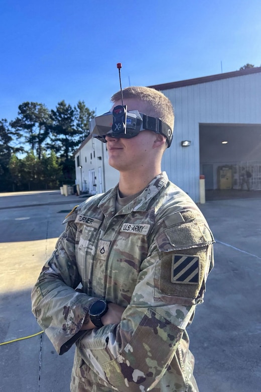 A soldier wearing a camouflage military uniform stands with his arms crossed while wearing first-person-view drone goggles.