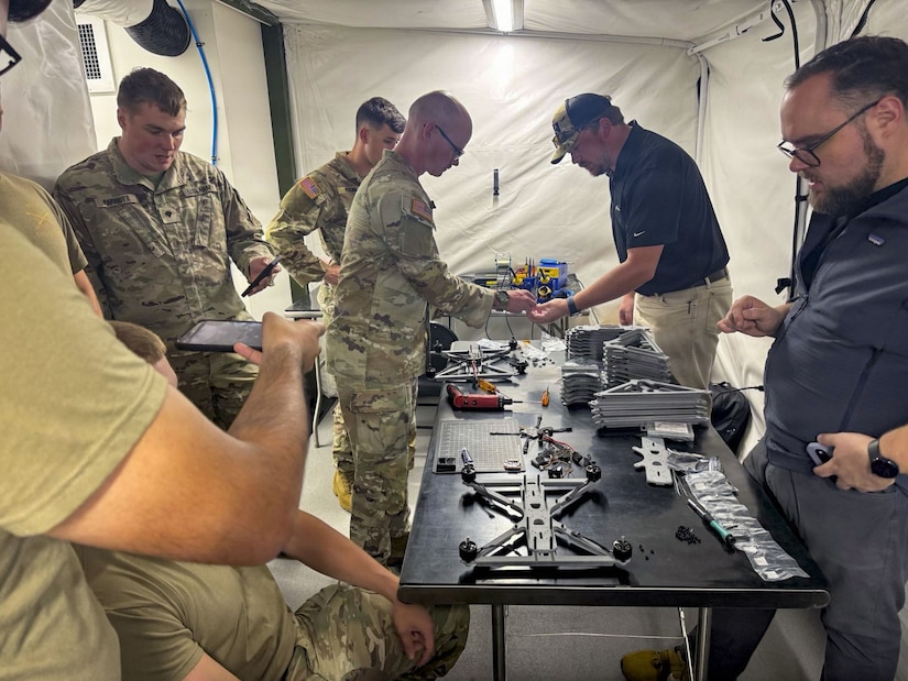 Men some dressed in camouflage military uniforms and others in civilian clothing stand around a table with drone parts.
