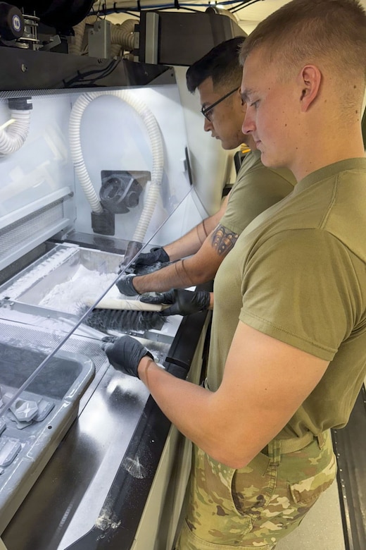 Two soldiers dressed in camouflage pants and t-shirt handle small drone parts.