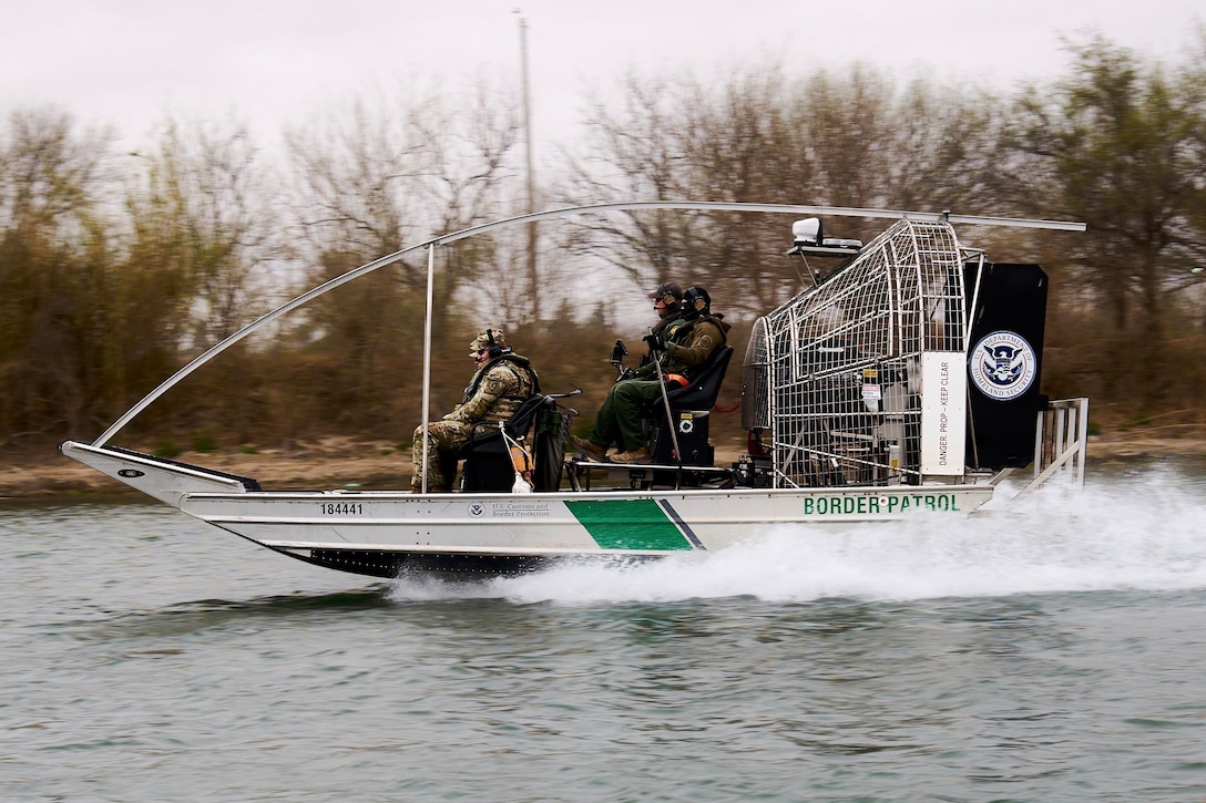 Soldiers and U.S. Customs and Border Protection agents ride in a small boat in a body of water on a gloomy day.