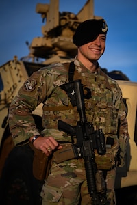 a security forces airmen leaning up against a combat vehicle smiling and laughing, beret on and gun across their chest, golden sunlight lighting