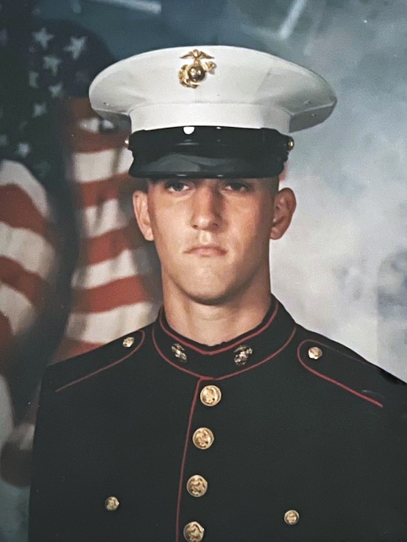 A Marine in a dress blue uniform poses for a photo with the American flag in the background.