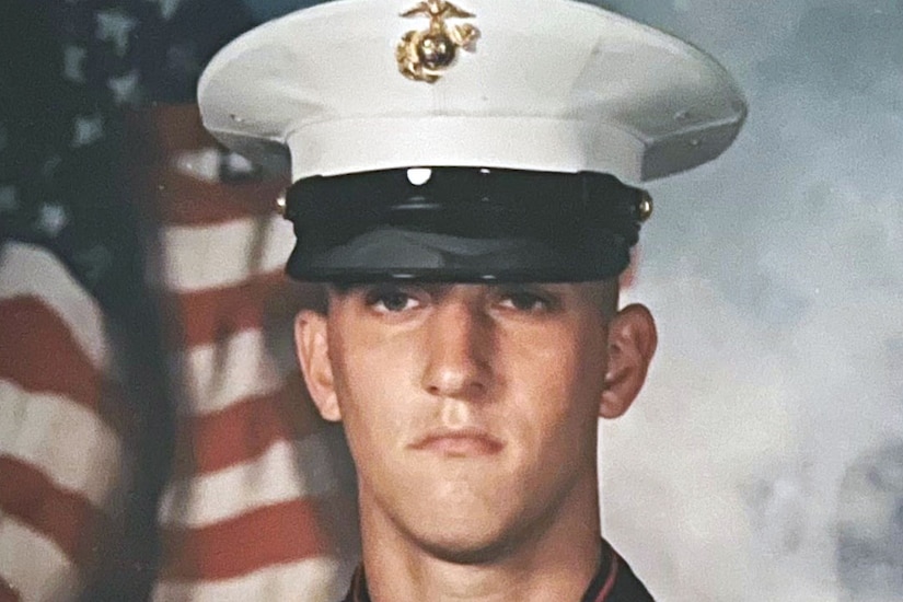 A Marine in a dress blue uniform poses for a photo with the American flag in the background.