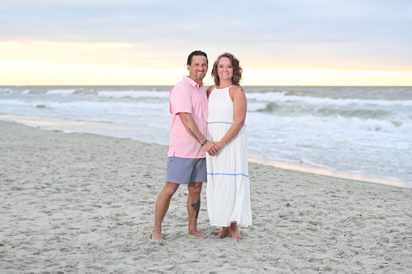 A man and a woman, in a slight embrace, pose for a photo on a beach, hands clasped.