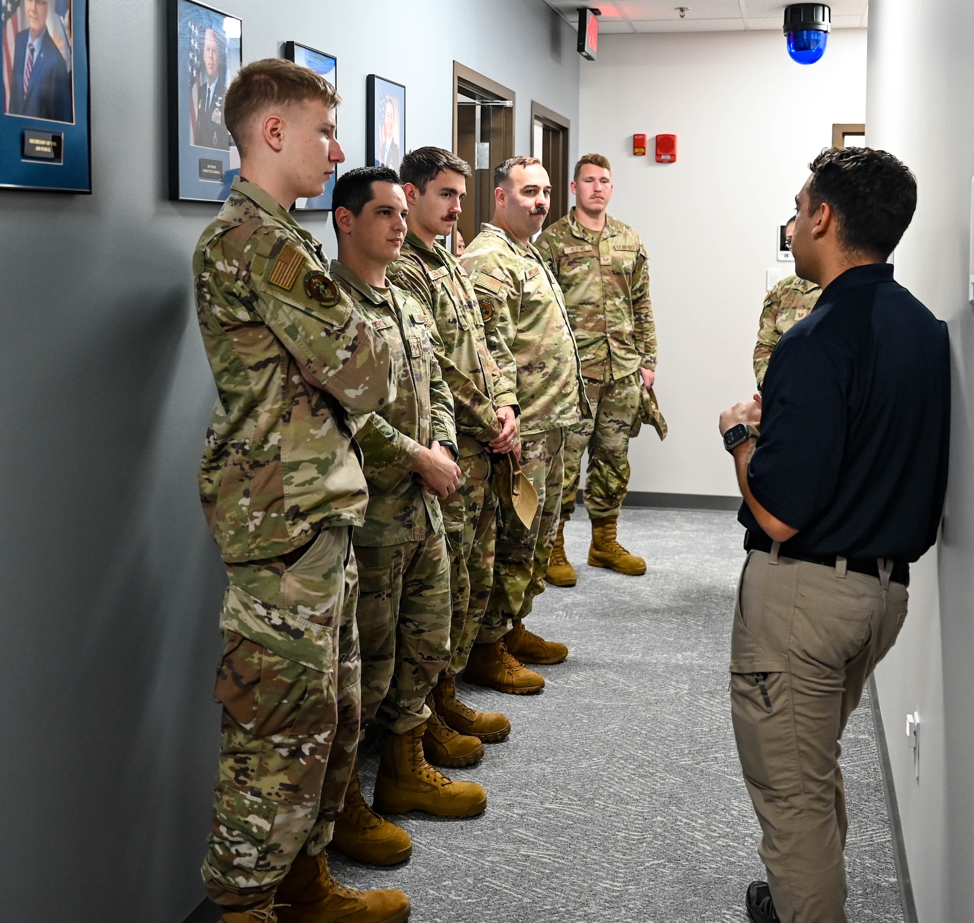 A group of military members in occupational camouflage pattern uniforms stand in a hallway listening to a man in khakis and a polo speak.