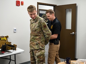 A military member in an occupational camouflage pattern uniform gets handcuffed by a man in khakis and a blue polo.