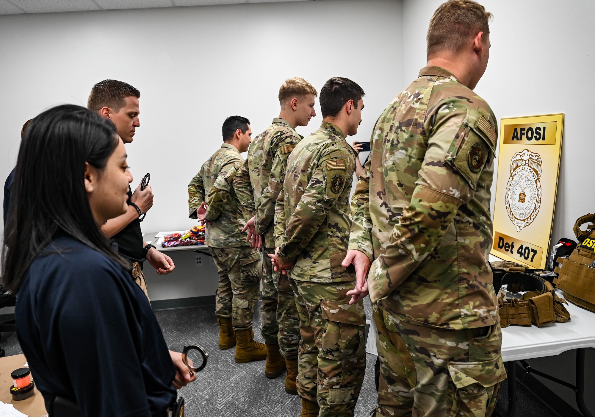 A group of military members in occupational camouflage pattern uniforms stand with their hands behind their backs waiting to be handcuffed by OSI agents in khakis and polos.