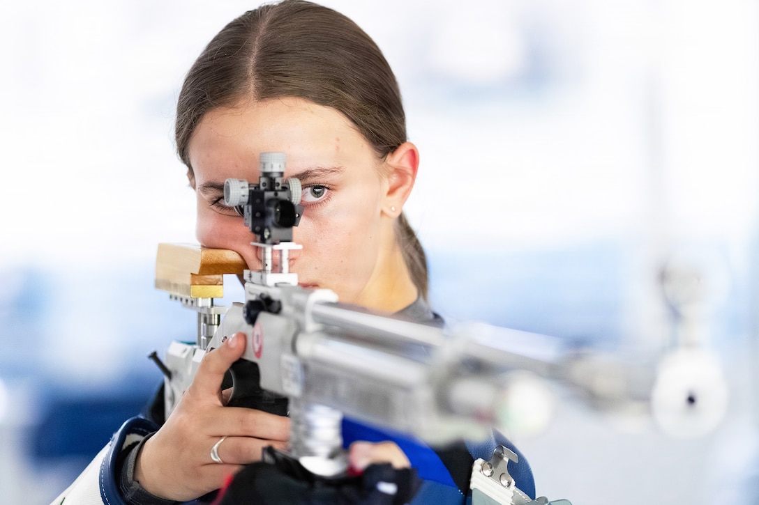 An Air Force Academy cadet holds and aims a silver rifle with one eye to a scope.