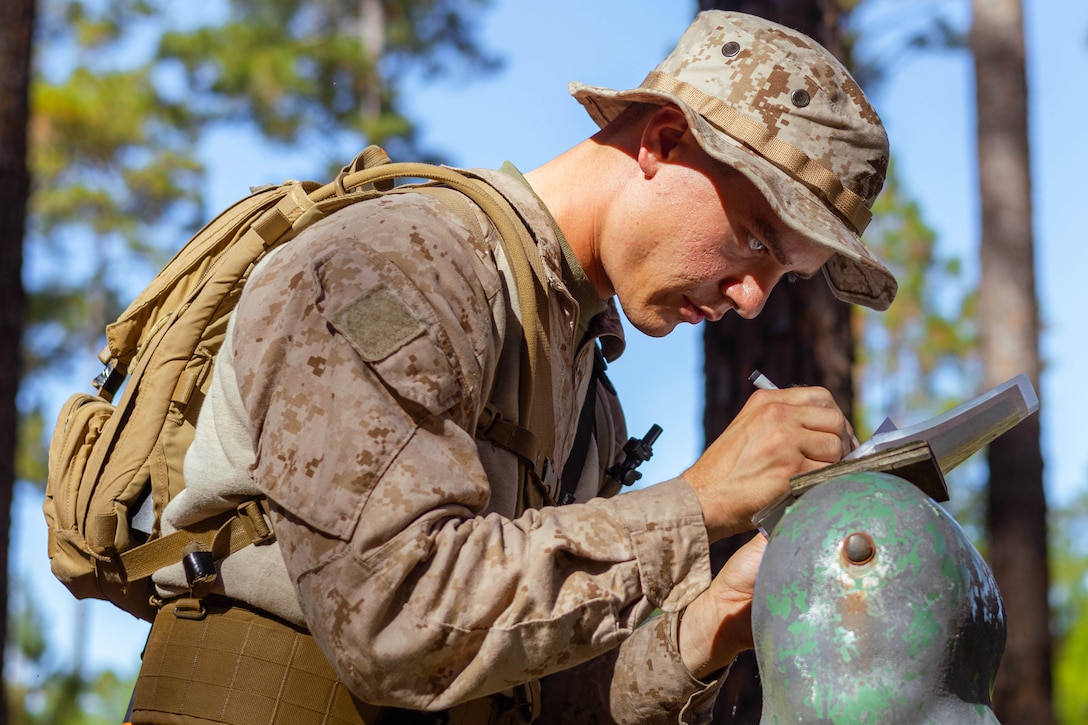 A Marine Corps recruit uses a pen to mark a point on a handheld tablet while standing outdoors during daytime, with trees in the background.