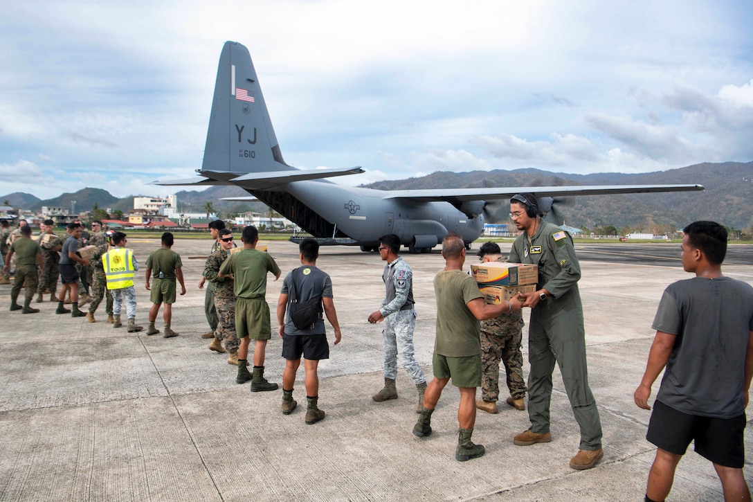 U.S. and Philippine military service members stand in a line on a runway and pass boxes to one another during daytime, with an airplane in the background and hills in the distance.