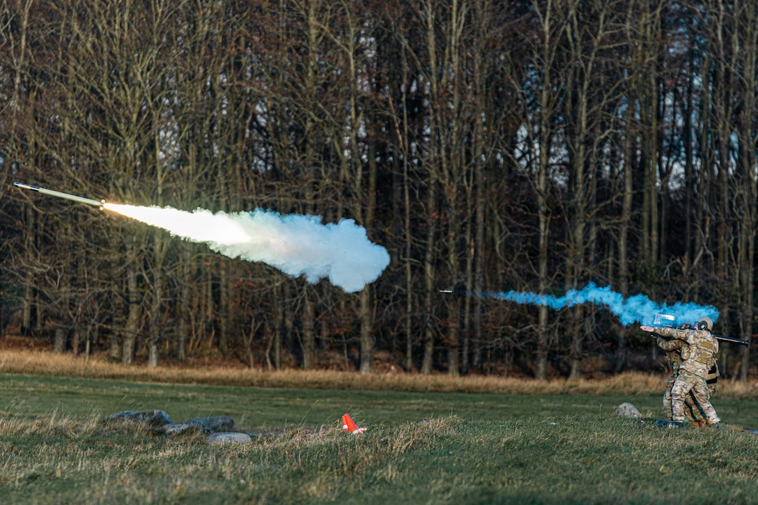 Two soldiers standing in a grassy field during daytime fire a missile into the air, leaving two trails of smoke, with woods in the background.