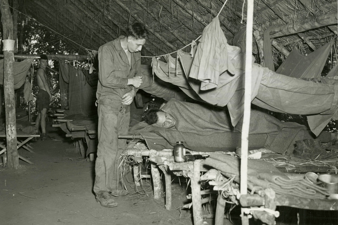 A man lies in a makeshift cot as another man stands over him. A third man looks at them from further away in the makeshift hospital.