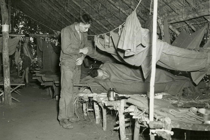 A man lies in a makeshift cot as another man stands over him. A third man looks at them from further away in the makeshift hospital.