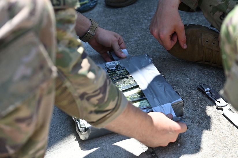 A person wearing a camouflage military uniform attaches duct tape to five rectangles of explosives.