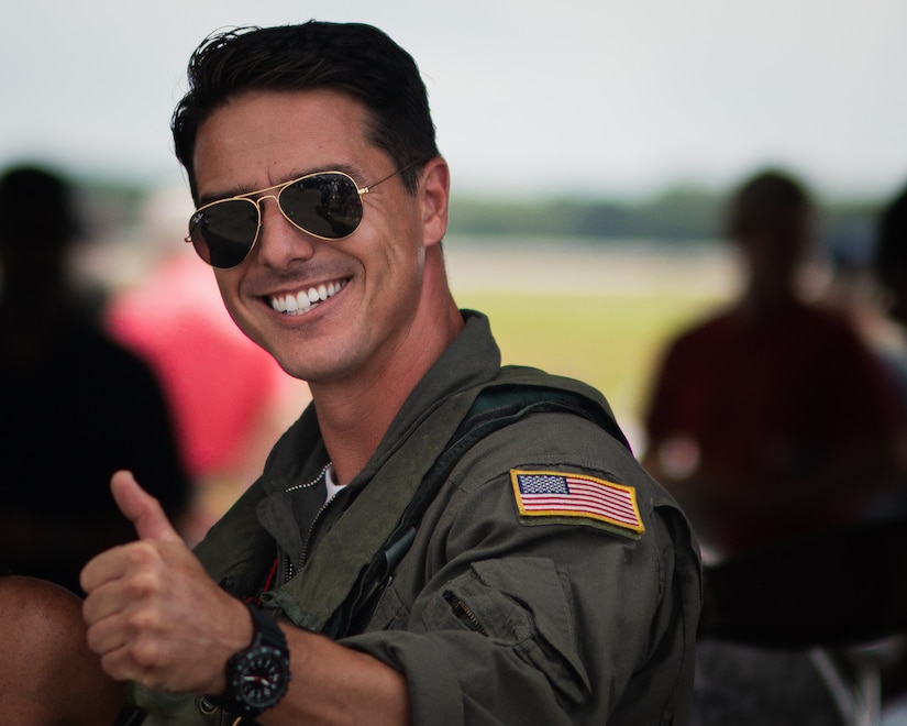 A man in aviator sunglasses and a flight suit smiles and gives a thumbs up.