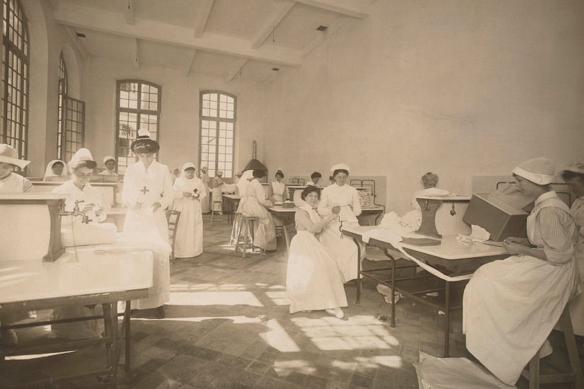Several women in nursing outfits sew long strips of bandages while seated at tables in a large room.