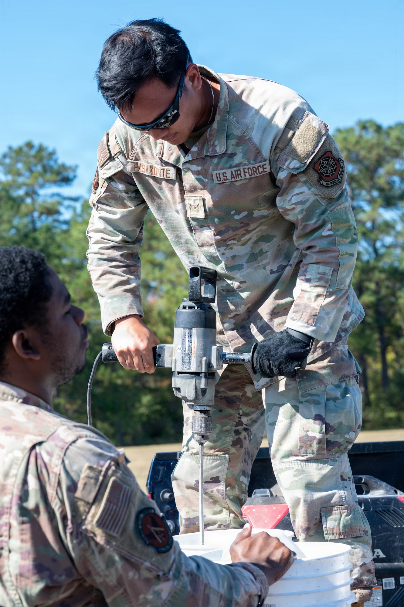 A photo of an Airman mixing concrete in a bucket and an Airman holding the bucket.