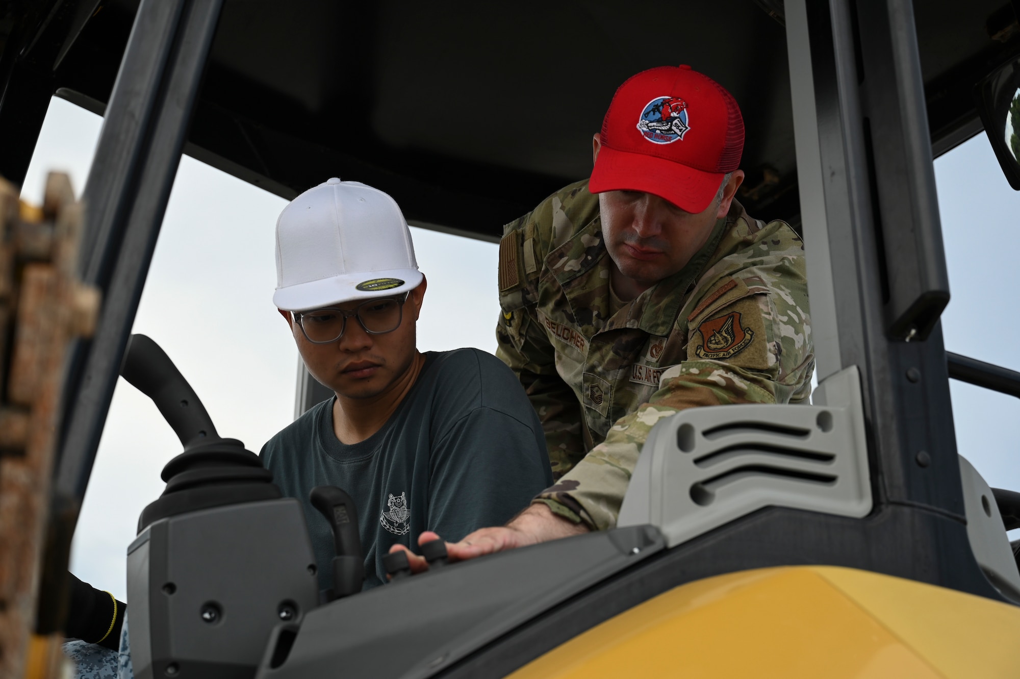 U.S. Air Force Master Sgt. with a red hat instructs and guides a foreign airman with a white hat on how to operate a vehicle.