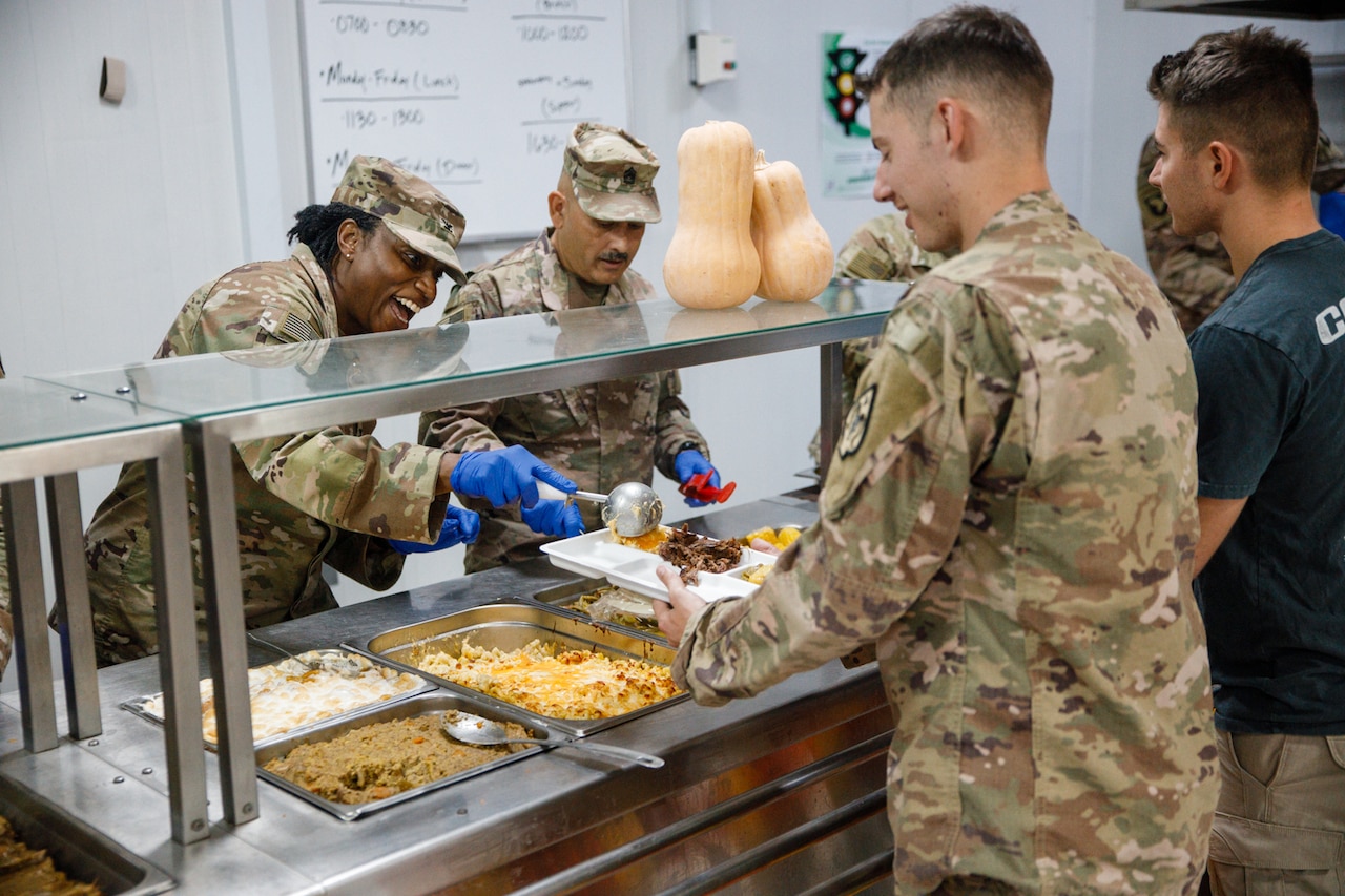 Service members in camouflage military uniforms serve food to other service members in similar attire that are standing in a cafeteria buffet line.