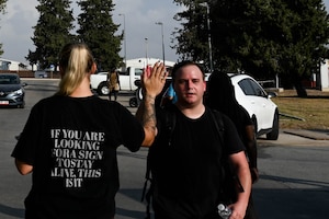 a blonde woman is high-fiving a man during a 5k ruck, and the back of the woman's shirt says "if you are looking for a sign to stay, this is it"