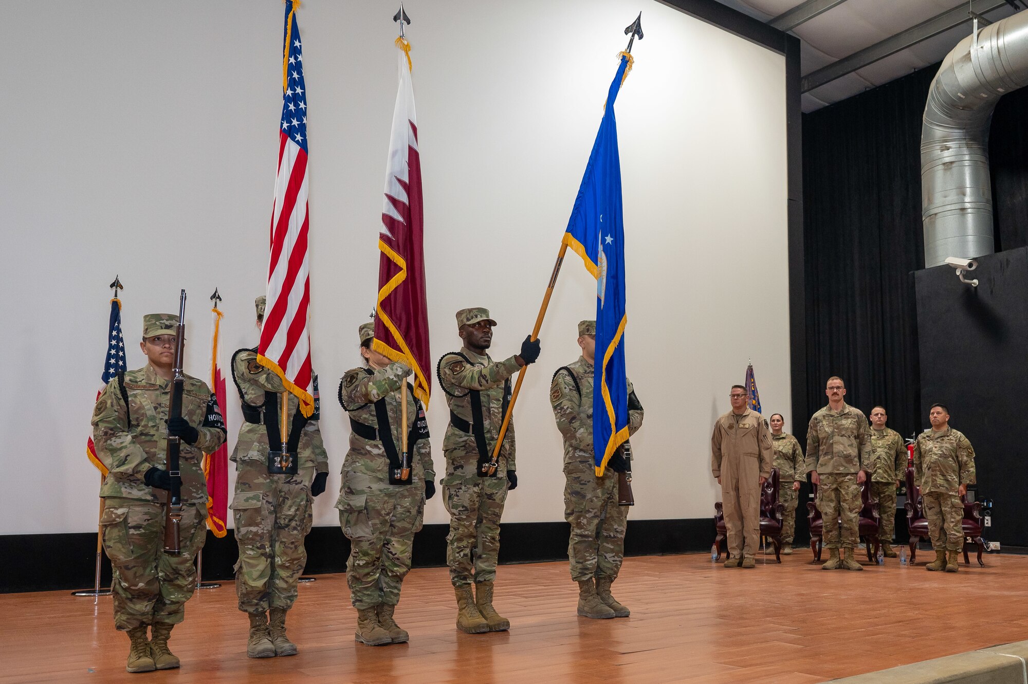 Five Airmen form an honor guard displaying the U.S., Qatar, and Air Force flags while people in the background stand at attention