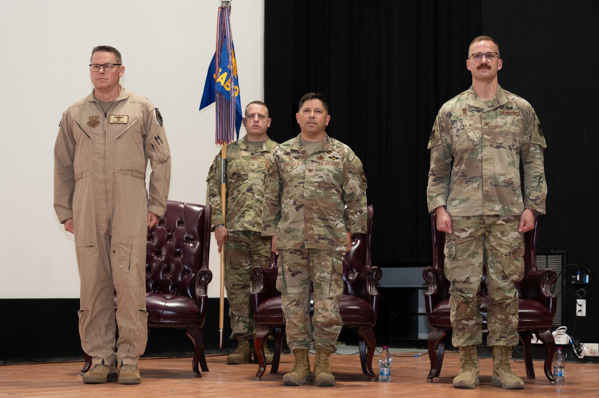 Three U.S. Air Force officers stand at attention on a stage in front of their chairs while a fourth Airman stands in the background holding a guidon