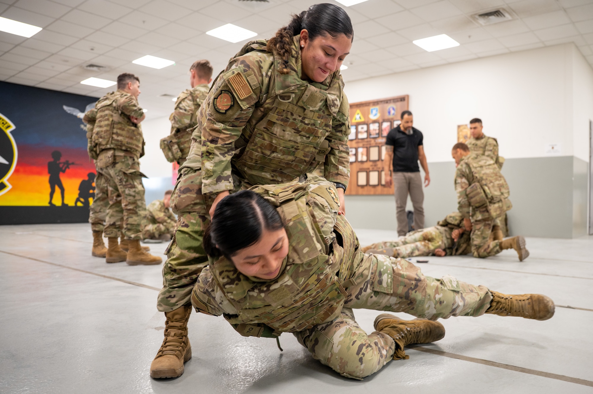 One Airman picks another up from a laying down position to kneeling. Other Airmen are in the background practicing takedown techniques.