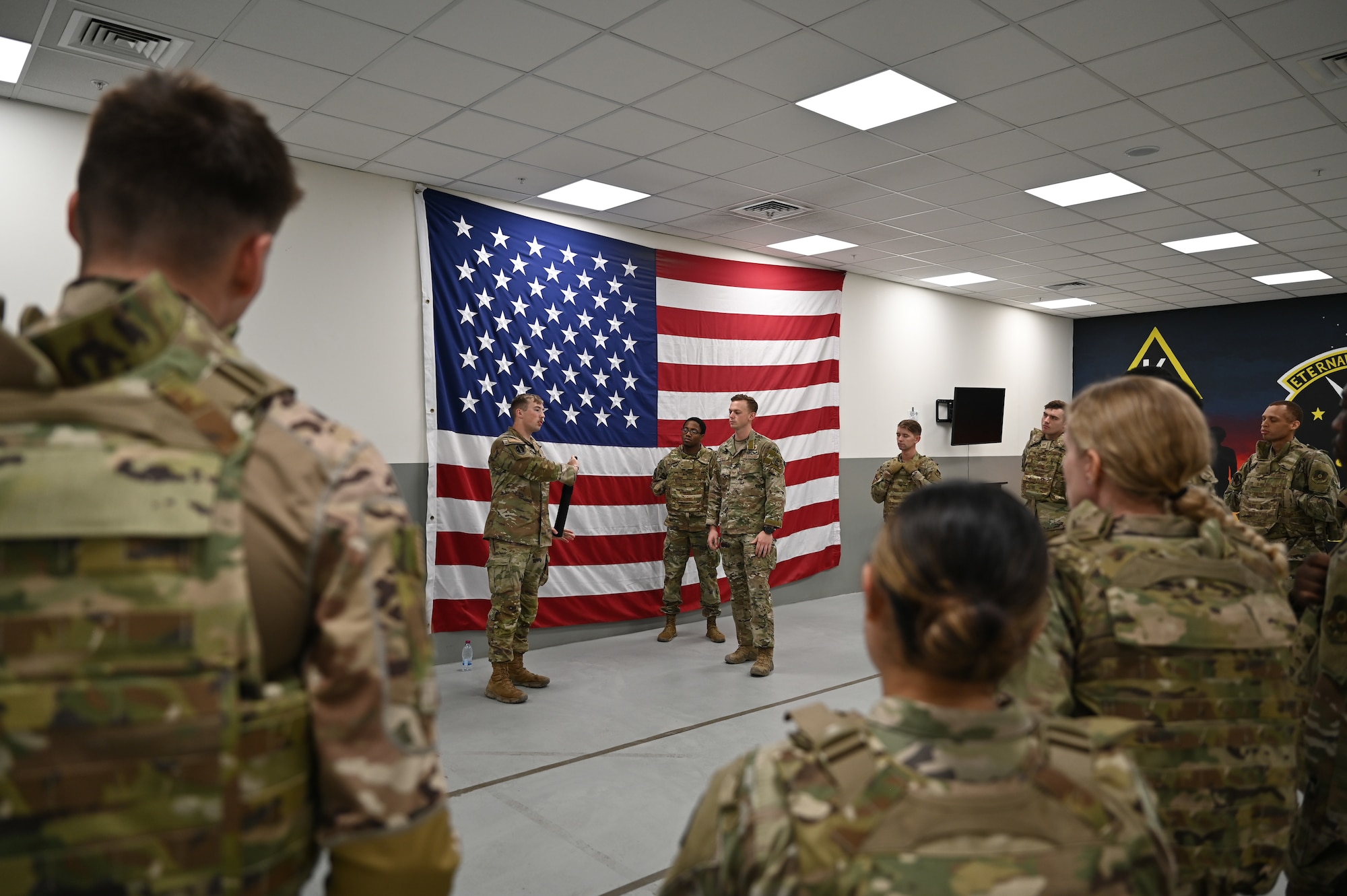 Two Airmen stand in front of a large American flag and speak to a crowd of Airmen who fill the foreground