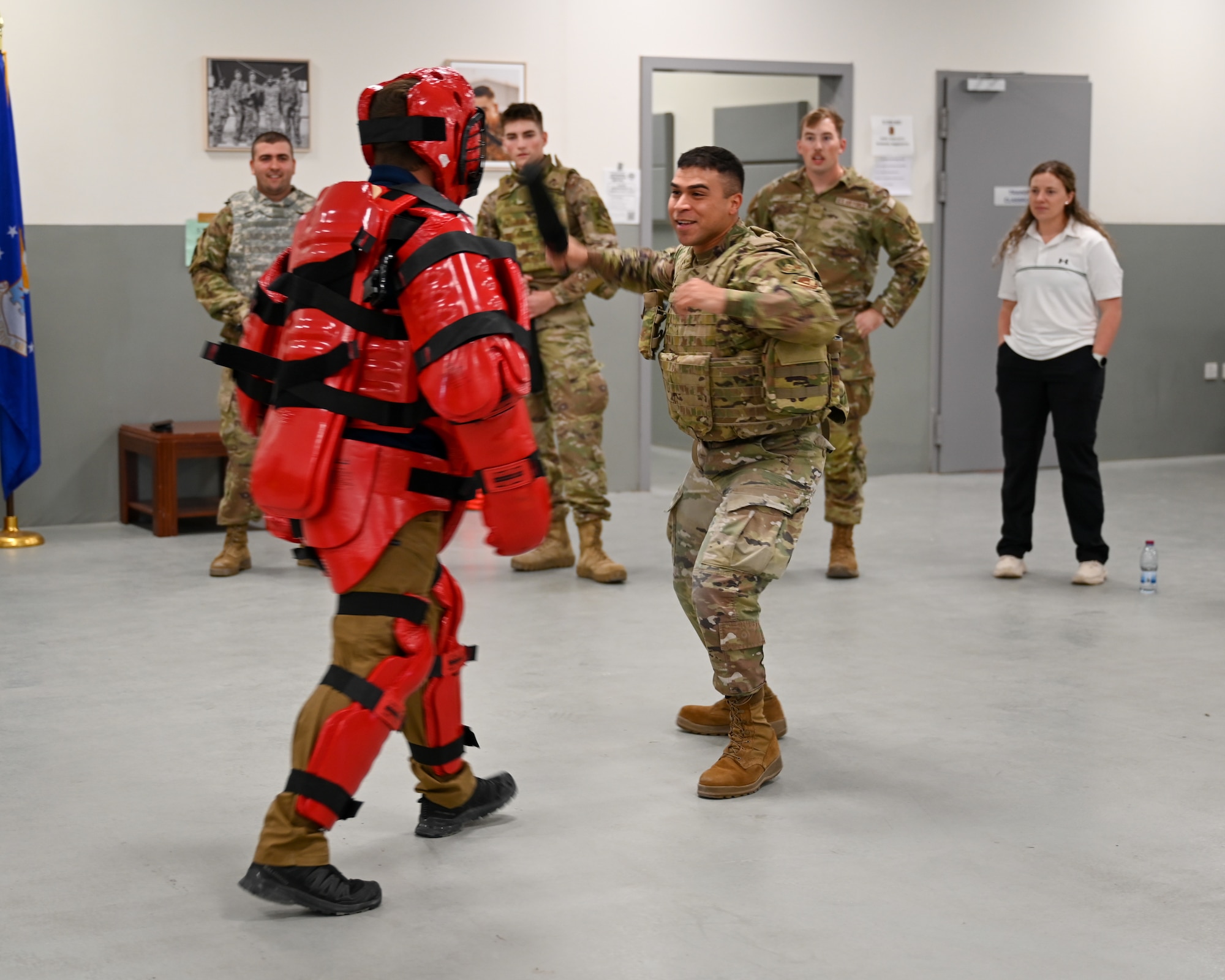 An Airman wields a padded baton at a man in a red protective suit as people in the background look on