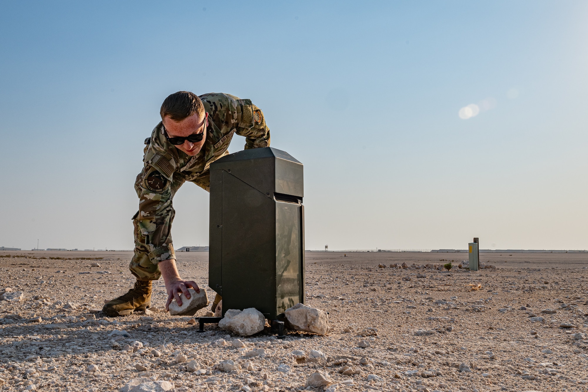 An Airman bends over with a rock in hand to place it on the base of a ceilometer