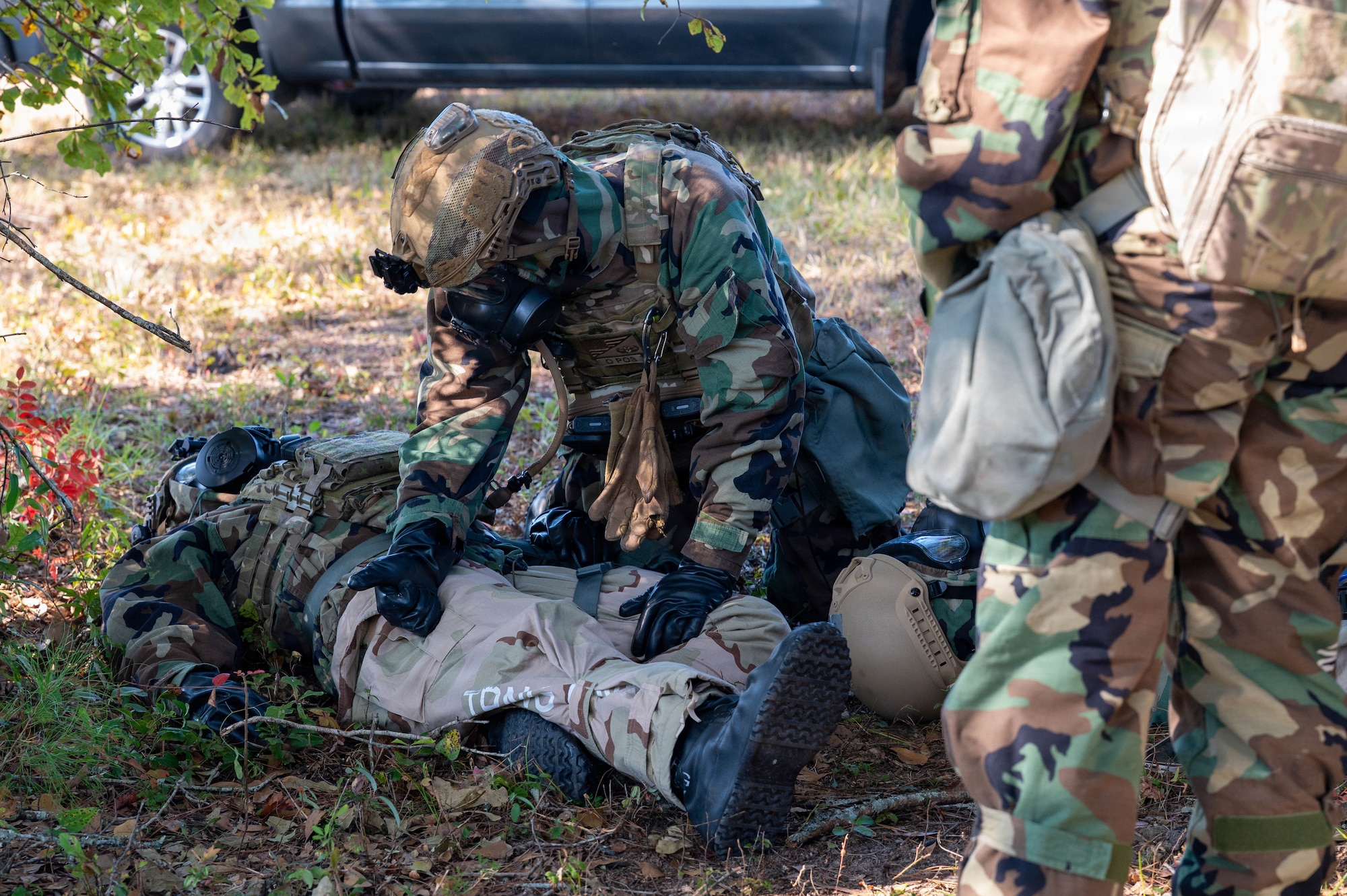 A photo of Airmen wearing gas masks and Mission Oriented Protective Posture gear engaging in a medical situation by laying on the ground, giving simulated medication and observing.