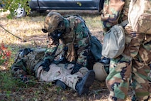 A photo of Airmen wearing gas masks and Mission Oriented Protective Posture gear engaging in a medical situation by laying on the ground, giving simulated medication and observing.