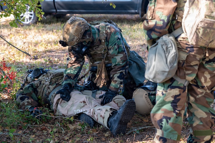 A photo of Airmen wearing gas masks and Mission Oriented Protective Posture gear engaging in a medical situation by laying on the ground, giving simulated medication and observing.