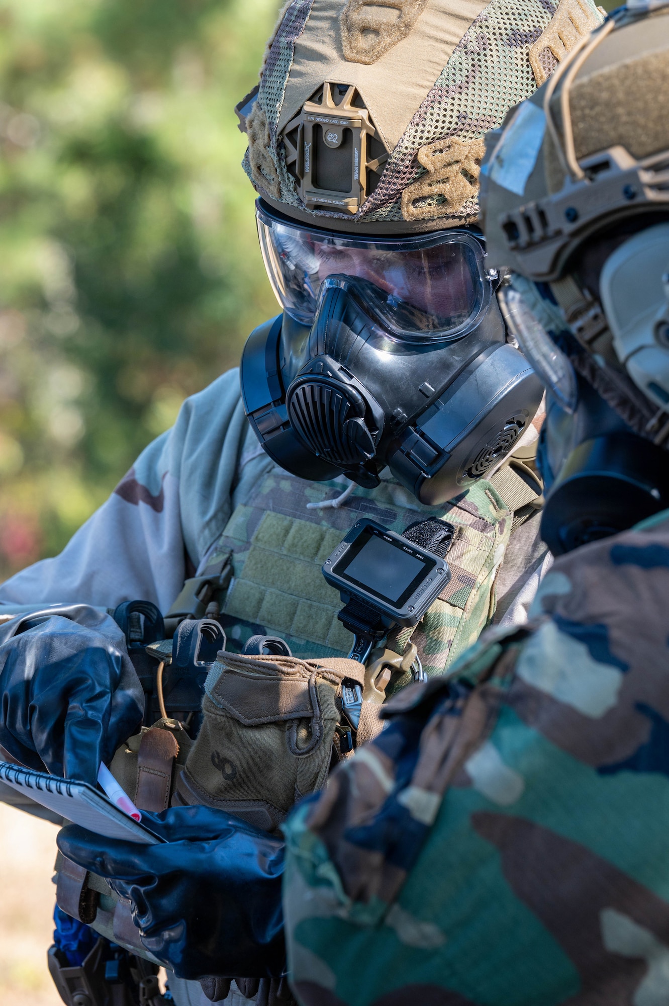 A photo of an Airman in a gas mask and Mission Oriented Protective Posture gear writes on a piece of paper.