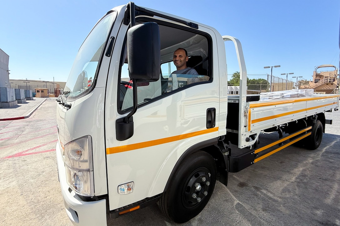 A man sits inside of a white cab-over-engine flatbed truck outside on a sunny, clear day.