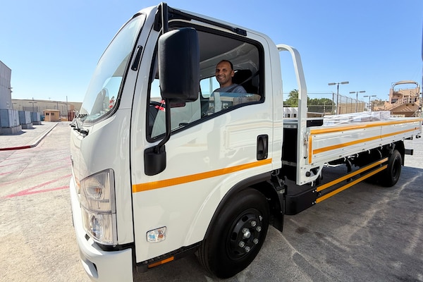 A man sits inside of a white cab-over-engine flatbed truck outside on a sunny, clear day.