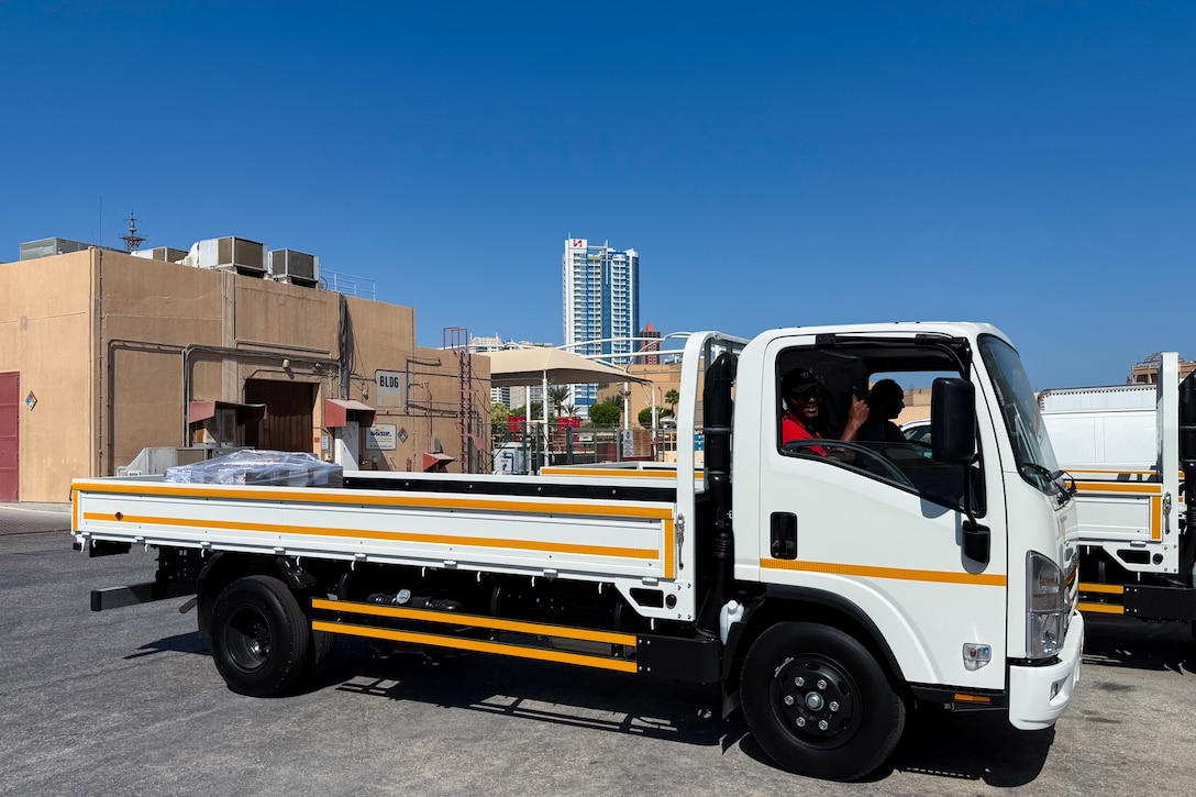 Two men sit inside of a cab-over-engine flatbed truck outside on a sunny, clear day.