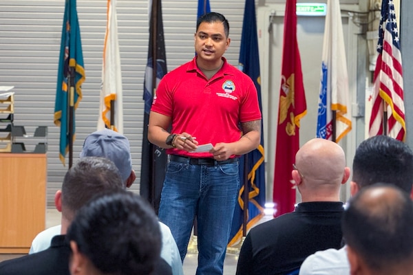 A man in a red polo shirt addresses a seated audience with military service flags behind him.